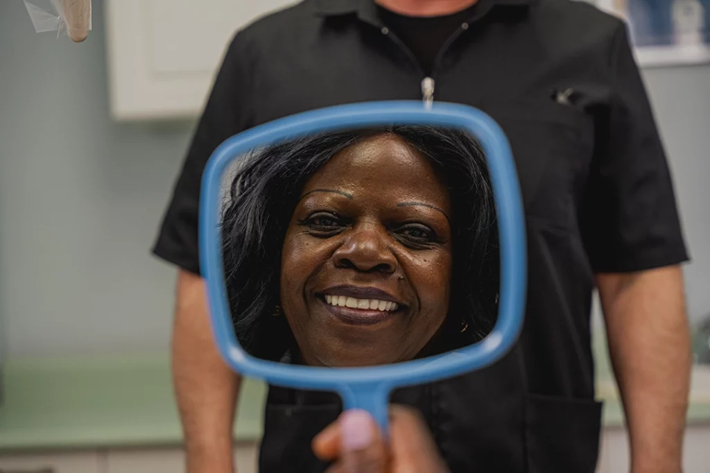 Patient of Battell Denture Clinic looking into a small, blue, handheld mirror smiling and looking at her new dentures.