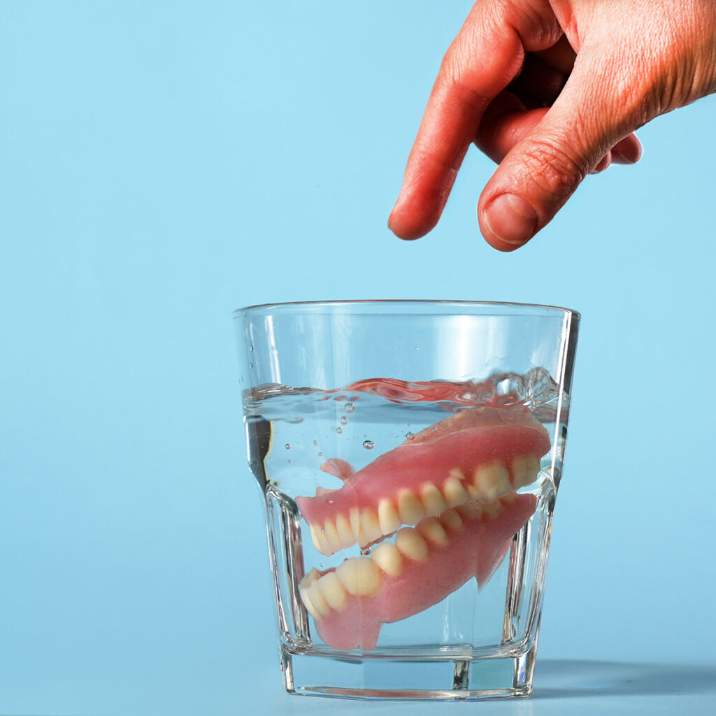 Close-up of a hand putting dentures in a glass of water and cleaning solution.