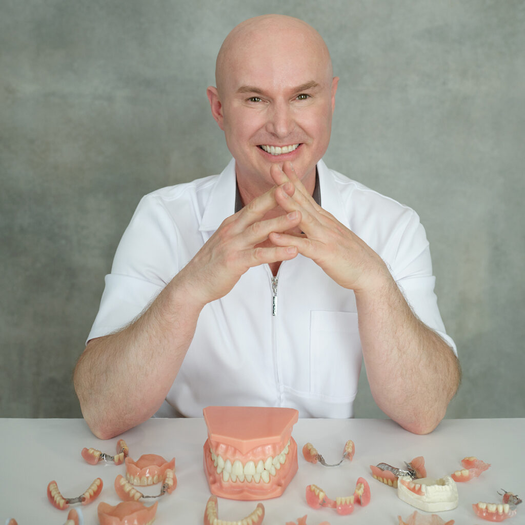 Michael Battell sitting at a desk, smiling, with a dozen of denture models laid out on the desk in front of him.
